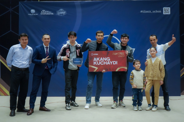 Gruppe von Menschen auf einer Bühne mit einer Tafel mit dem Text 'Akang Kuchaydi - Zakovat Quiz', mit einem Banner, einem Barcode und Lichtern im Hintergrund.