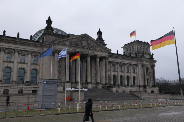 Eine Person, die vor dem Reichstaggebäude in Berlin, Deutschland, mit seinen architektonischen Details, Flaggen, Treppen, Geländern, Bäumen und klarem Himmel spaziert.