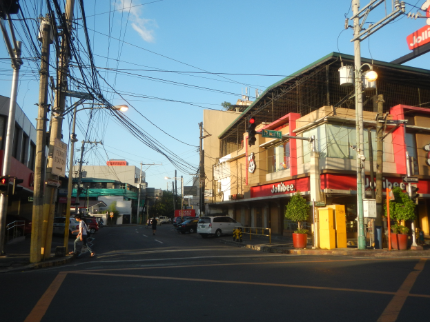 Eine Stadtstraße mit Autos, Fußgängern, Strommasten, Verkehrsampeln, Gebäuden mit Schildern, Topfpflanzen und einer bewölkten Himmel.