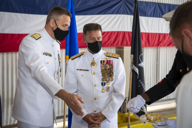 Zwei Männer in weißen Uniformen und Masken stehen nebeneinander, einer hält ein Tablett mit einem Lebensmittel, mit Flaggen und einem Vorhang im Hintergrund.