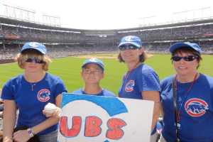 Drei Frauen in Kappen und Brillen, ein Junge in Brille und Kappe mit einem Ball und einer Tafel, stehend in einem Stadion mit Publikum und Feld dahinter, unter einem Himmel mit einigen Lichtern.
