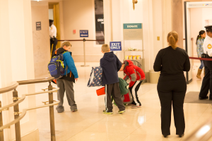 Eine Gruppe von Menschen steht in einem Flur mit einem Geländer auf der linken Seite, Deckenleuchten und Tafeln mit Text im Hintergrund, was darauf hindeutet, dass es der erste Schultag im U.S. Capitol ist.