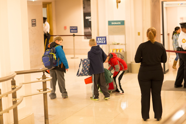 Eine Gruppe von Menschen steht in einem Flur mit einem Geländer auf der linken Seite, Deckenleuchten und Tafeln mit Text im Hintergrund, was darauf hindeutet, dass es der erste Schultag im U.S. Capitol ist.