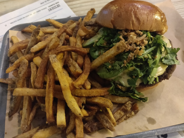 Ein Tablett mit einem Hamburger, der mit Salat, Tomate und Zwiebeln belegt ist und goldenen, knusprigen Pommes frites, das auf einem Tisch mit einem Blatt daneben steht.