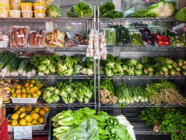Ein Gang in einem Supermarkt mit frischem Gemüse und Obst, verpackten Artikeln in Plastik, Kartons, eine Texttafel und ein Glasfenster im Hintergrund.