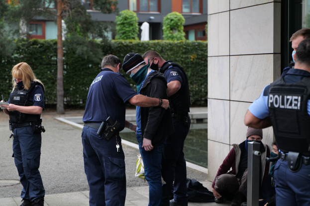 Eine Gruppe von Polizisten steht vor einem Gebäude, während eine Frau mit einem Buch und einem Stift ihnen gegenübersteht, mit sitzenden und stehenden Personen in der Nähe und Grünflächen und Gebäuden im Hintergrund.