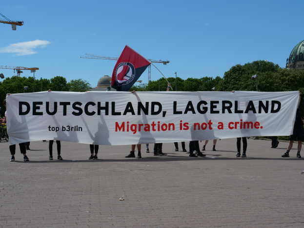 Eine Gruppe von Menschen, die draußen vor einem Banner mit der Aufschrift "Deutschland, Lagerland Migration ist kein Verbrechen" und einer Flagge stehen, mit Straßenlaternen, Bäumen, Kränen, einem Gebäude und einem bewölkten Himmel im Hintergrund.