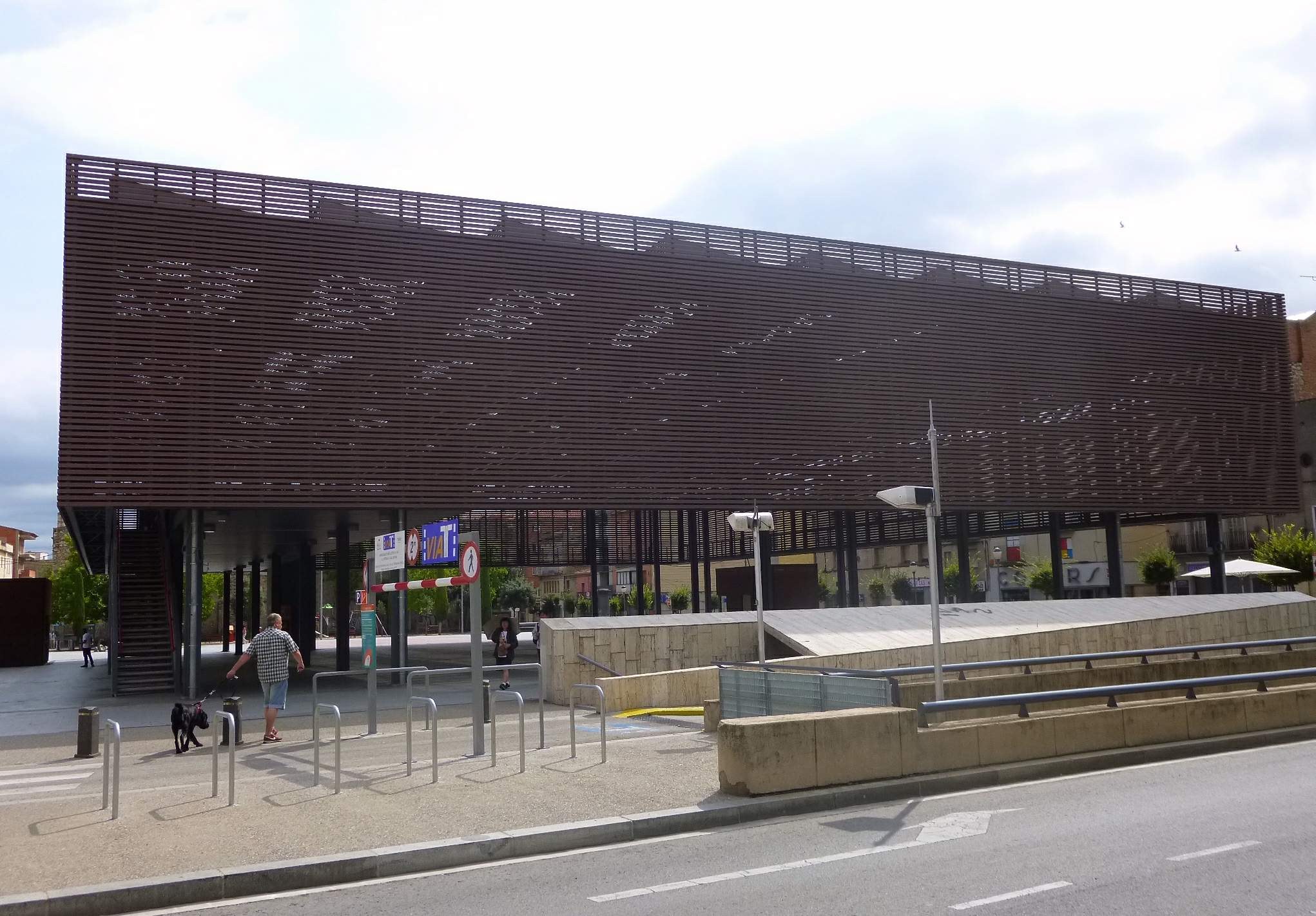 Großes modernes Gebäude mit zahlreichen Fenstern, das den neuen Eingang zur Stadtmitte bildet, umgeben von Straßeninfrastruktur, Fahrzeugen, Fußgängern und Bäumen unter einem bewölkten Himmel.