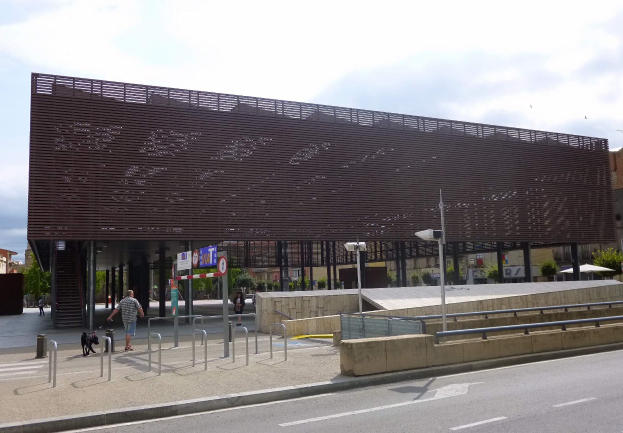 Großes modernes Gebäude mit zahlreichen Fenstern, das den neuen Eingang zur Stadtmitte bildet, umgeben von Straßeninfrastruktur, Fahrzeugen, Fußgängern und Bäumen unter einem bewölkten Himmel.