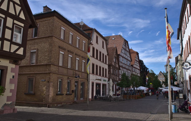Headsteinstraße in Rothenburg ob der Tauber, Deutschland, gesäumt von Gebäuden mit Fenstern, Bäumen und Fahnenmasten, mit einigen Menschen und Fahrzeugen auf der Straße unter einem bewölkten Himmel.