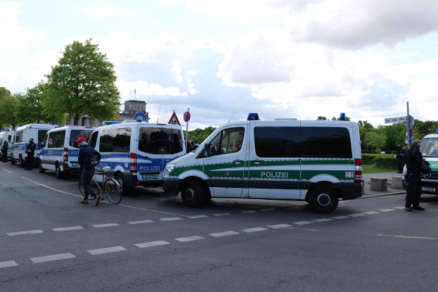 Eine Gruppe von Polizeifahrzeugen, die entlang einer Straße geparkt sind, mit einem Radfahrer im Vordergrund, Bäumen, Pfosten, Schildern, Gebäuden und einem bewölkten Himmel im Hintergrund.