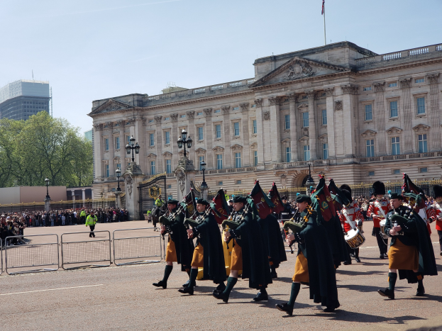 Gruppe von Absolventen in Roben und Hüten mit Musikinstrumenten vor einem Gebäude mit Metallzaun, Straßenlaterne, Bäumen und bewölktem Himmel.