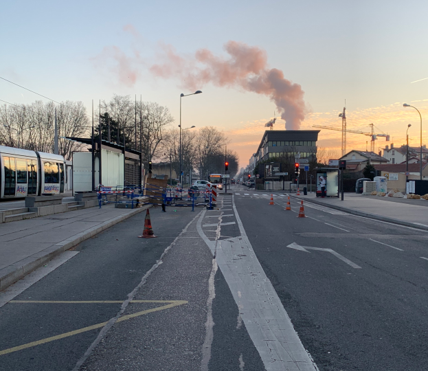 Eine Straßenansicht mit Absperrbaken, einem Bus, Laternenpfählen, Bäumen, Gebäuden, Kränen und Rauch, der aus einem entfernten Gebäude aufsteigt, unter einem klaren Himmel.