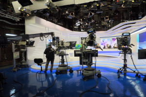 Ein Mann steht vor einem Fernseher und hält ein Mikrofon, umgeben von Kameras, einem Stuhl, einem Bildschirm, einer Wand und einer Decke mit Scheinwerfern, was auf eine Live-Übertragung hindeutet.