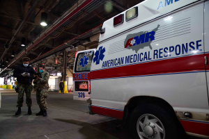 Ein Rettungswagen der American Medical Response (AMR) steht in einem Gebäude geparkt, mit zwei maskierten Personen davor, einem Fahrzeug auf der linken Seite und mehreren Personen, Lichtern und Rohren im Hintergrund.