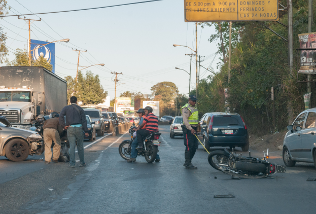 Eine Gruppe von Menschen umringt ein verunglücktes Motorrad am Straßenrand mit mehreren Fahrzeugen, darunter ein Lastwagen, und einem Hintergrund aus Bäumen, Polen, Lampen und Schildern unter dem Himmel.