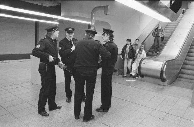 Schwarzes und weißes Bild von Polizisten, die in einer U-Bahn-Station mit einer Rolltreppe und Deckenbeleuchtung im Hintergrund stehen.