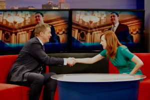 Ein Mann in einem schwarzen Blazer und weißem Hemd gibt einer Frau in einem grünen Kleid die Hand, vor einer roten Studiosofa stehend, mit mehreren Bildschirmen an der Wand im Hintergrund.