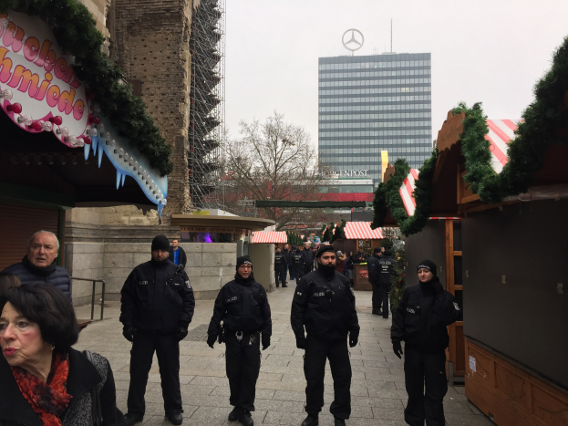 Polizeibeamte in schwarzen Uniformen stehen vor einem geschmückten Weihnachtsmarkt in Berlin, mit festlichen Dekorationen, Gebäuden, Bäumen und einem klaren blauen Himmel im Hintergrund; ein Schild mit Text ist links sichtbar.