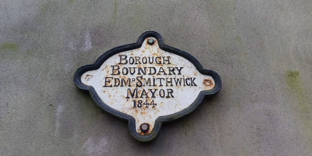 Metallplatte an der Seite eines Gebäudes, die die Bezirksgrenze markiert, mit "Bezirksgrenze Edm Smithwick Bürgermeister" in fetten weißen Buchstaben auf einem schwarzen Hintergrund.