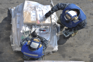 Zwei Männer in blauen Uniformen und Helmen arbeiten an einem großen Drohnenmodell, halten verschiedene Gegenstände und haben eine Plastikabdeckung auf dem Boden.