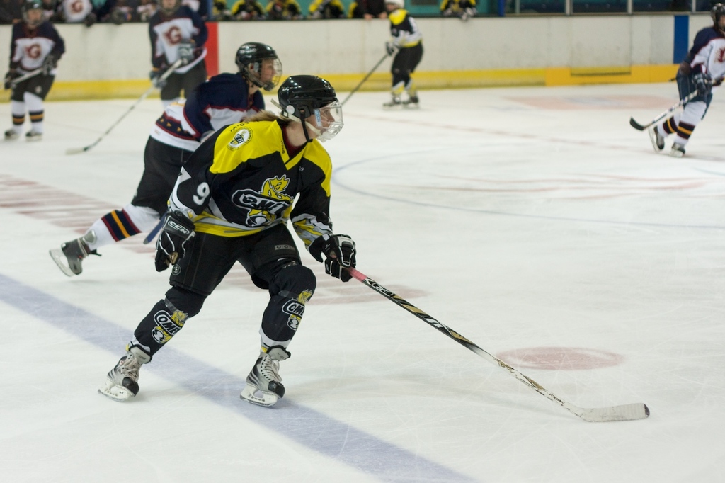 Menschen beim Schneehockey mit Hockeystöcken und Helmen, mit anderen und einer Wand im Hintergrund.