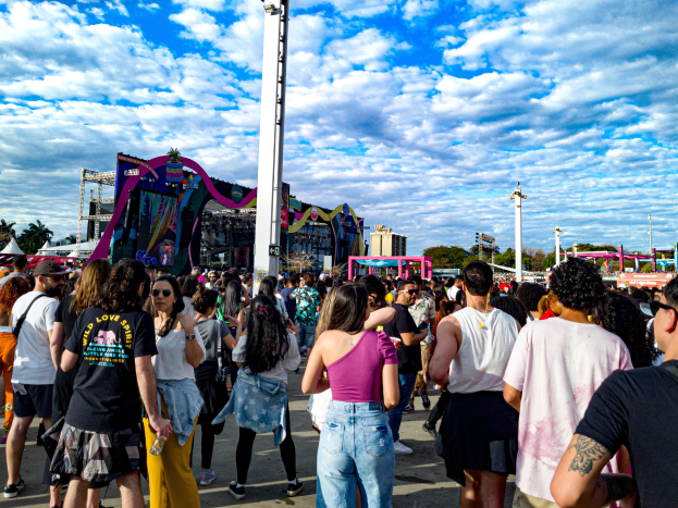 Eine Menschenmenge, einige mit Mützen und Schutzbrillen, steht vor einem Fasching mit Pfählen, Bäumen, Gebäuden und einem bewölkten Himmel im Hintergrund.