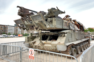 Ein gepanzerter Mannschaftswagen steht vor einem Metallzaun mit einem daran befestigten Schild, mit Fahrzeugen, Bäumen, Gebäuden und einem klaren blauen Himmel im Hintergrund.
