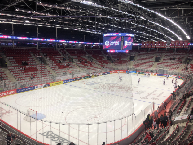 Eishockeyspiel in einer großen Arena zwischen den St. Louis Blues und den Detroit Red Wings, mit Zuschauern auf den Sitzen und Stehplätzen in der Nähe des Spielfelds, umgeben von einem Zaun und Metallrahmen, unter einem Dach mit Deckenbeleuchtung und einem Bildschirm.