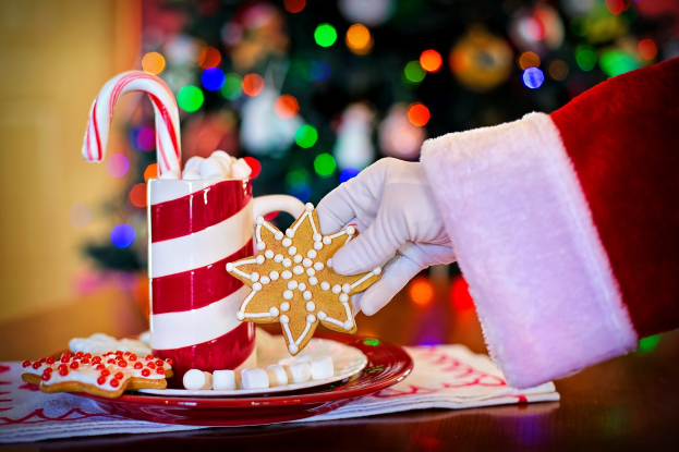 Eine Person mit einem Weihnachtsmannhut hält einen Becher heiße Schokolade und einen Zuckerstange in der Hand und hat einen Teller mit Keksen und einem Tuch auf dem Tisch vor sich, vor einem Weihnachtsbaum und einer Wand.