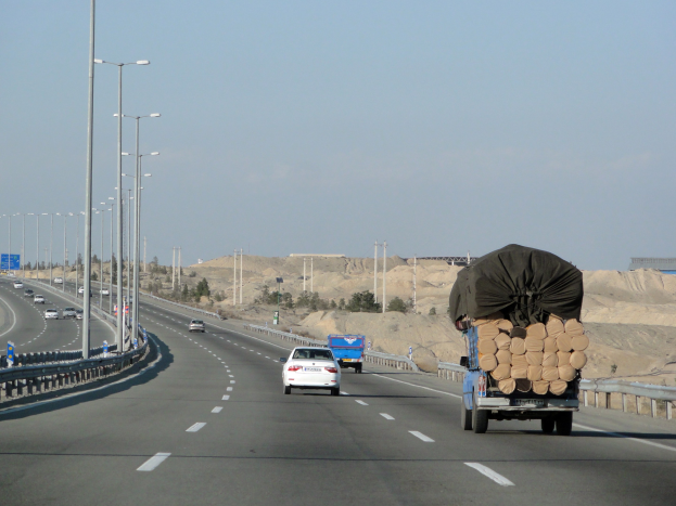 Ein Holzlastwagen mit einer großen Ladung Holz fährt auf einer Autobahn mit Schutzplanken, Laternen, Schildern, Bäumen und Sand, mit Hügeln und einem klaren blauen Himmel im Hintergrund.