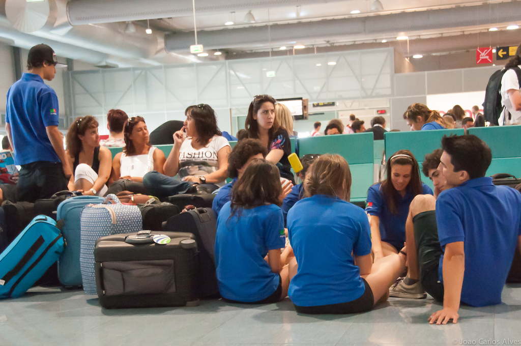 Ein belebtes Flughafeninnere mit vielen Menschen, die auf Flüge warten, Gepäck auf der linken Seite und Glastüren mit Richtungsschildern im Hintergrund.