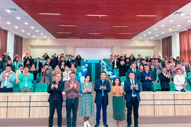 Eine Gruppe von Menschen steht vor einer Menge, einige klatschen, mit Stühlen, Tischen, Ballons, Vorhängen und einer Wand im Hintergrund sowie Lampen an der Decke, bei der Eröffnungsfeier einer neuen Schule.