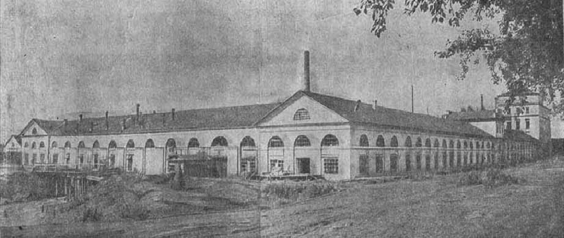Schwarzes und weißes Foto eines großen mehrstöckigen Gebäudes mit zahlreichen Fenstern, umgeben von Bäumen und Grün, unter einem klaren Himmel, mit dem Text "1892 – das erste Fabrikgebäude in der Stadt" unten.