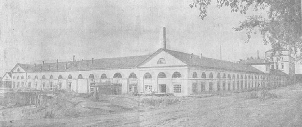 Schwarzes und weißes Foto eines großen mehrstöckigen Gebäudes mit zahlreichen Fenstern, umgeben von Bäumen und Grün, unter einem klaren Himmel, mit dem Text "1892 – das erste Fabrikgebäude in der Stadt" unten.