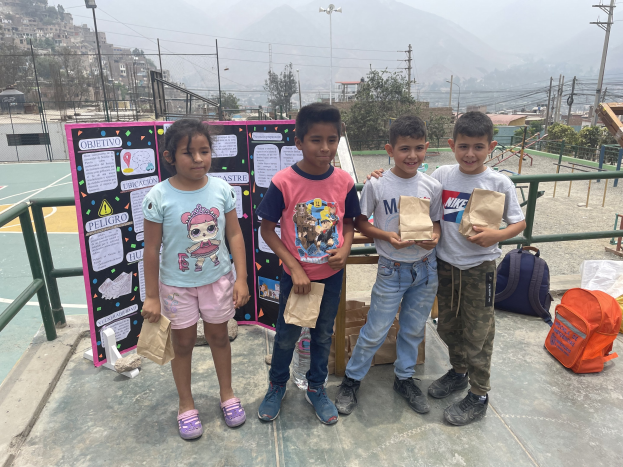 Gruppe von Kindern auf einem Basketballfeld mit Papierbeuteln in den Händen, Beutel und Gegenstände auf der rechten Seite und eine Tafel mit Text auf der linken Seite, vor einem Hintergrund aus Zäunen, Pfählen, Straßenlaternen, Strommasten, Bäumen, Gebäuden, Hügeln und Himmel.