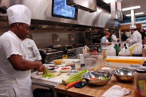 Eine Gruppe von Köchen in einer Küche beim Zubereiten von Speisen, mit einem Koch im Vordergrund in einer weißen Schürze und Mütze, der ein Messer hält und von Utensilien, Schüsseln, Gemüse, einem Handy, Flaschen und anderen Gegenständen umgeben ist, während im Hintergrund ein Fernseher und Deckenleuchten zu sehen sind.