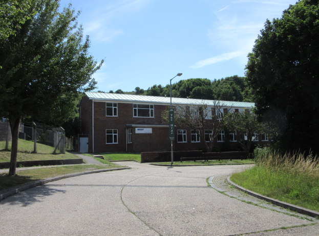 Ein Schulgebäude mit Fenstern, ein Straßenschild, ein Schild, eine Gruppe von Bäumen, Gras, ein Zaun und ein bewölkter Himmel, das in der Mitte eines Parks umgeben von saftigem Gras liegt.