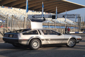 Ein silberner DeLorean DMC-12 Wagen befindet sich auf einer Straße, umgeben von Menschen, Fahnen, Schildern, Geländern, Stühlen und einem Schuppen, mit Bäumen und einem klaren blauen Himmel im Hintergrund.