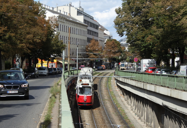 Rote und weiße Straßenbahn auf Schienen neben einer Stadtstraße mit Fahrzeugen, Gebäuden, Bäumen und Strommasten im Hintergrund.