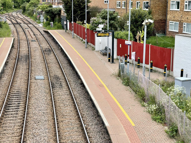Ein Bahnsteig mit zwei parallelen Gleisen, eine Plattform auf der linken Seite, Bäume und Gebäude im Hintergrund und ein Zaun auf der rechten Seite.