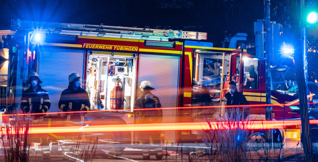 Feuerwehrleute in Schutzausrüstung stehen vor einem beleuchteten Feuerwehrauto in der Nacht, mit Pflanzen im Vordergrund, einem Verkehrszeichen auf der rechten Seite und Bäumen im Hintergrund.