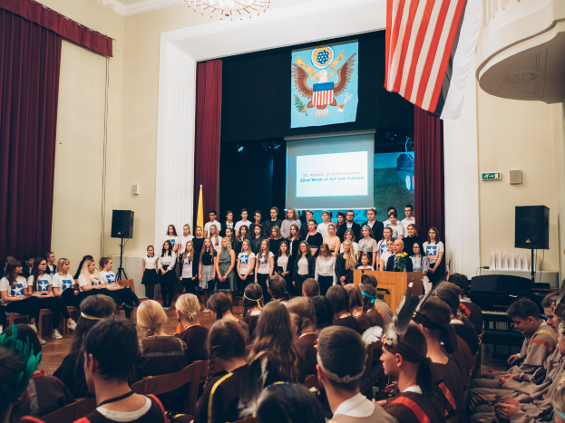 Eine Gruppe von Menschen steht an einem Podium mit einem Mikrofon während eines 2. Wochenende von Kunst und Kultur, flankiert von Fahnen und Lautsprechern, gegenüber einer sitzenden Menge mit einem Bildschirm und einer Deckenleuchte im Hintergrund.