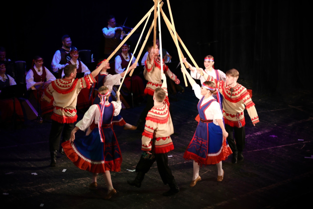 Eine Gruppe von Menschen in farbenfrohen Kostümen tanzt um einen Maibaum, einige halten Holzstöcke, mit Musikern im Hintergrund.