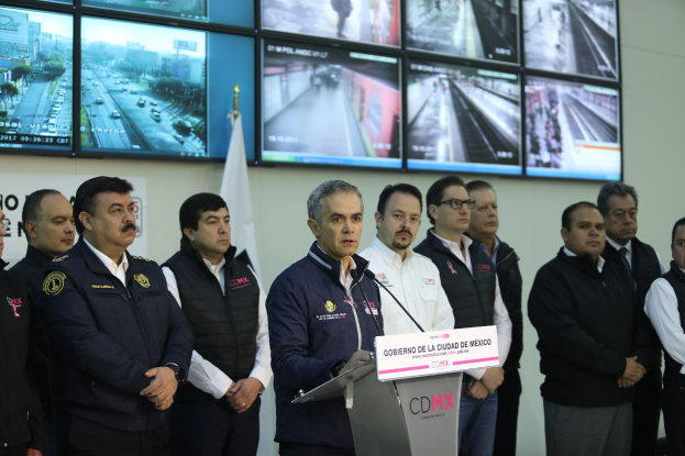 Gruppe von Männern, die vor einem Podium mit einem Mikrofon und einer Tafel mit der Aufschrift "Mexikanische Zoll- und Grenzschutzbehörde" stehen, mit Fernsehern und einer Flagge an der Wand dahinter.
