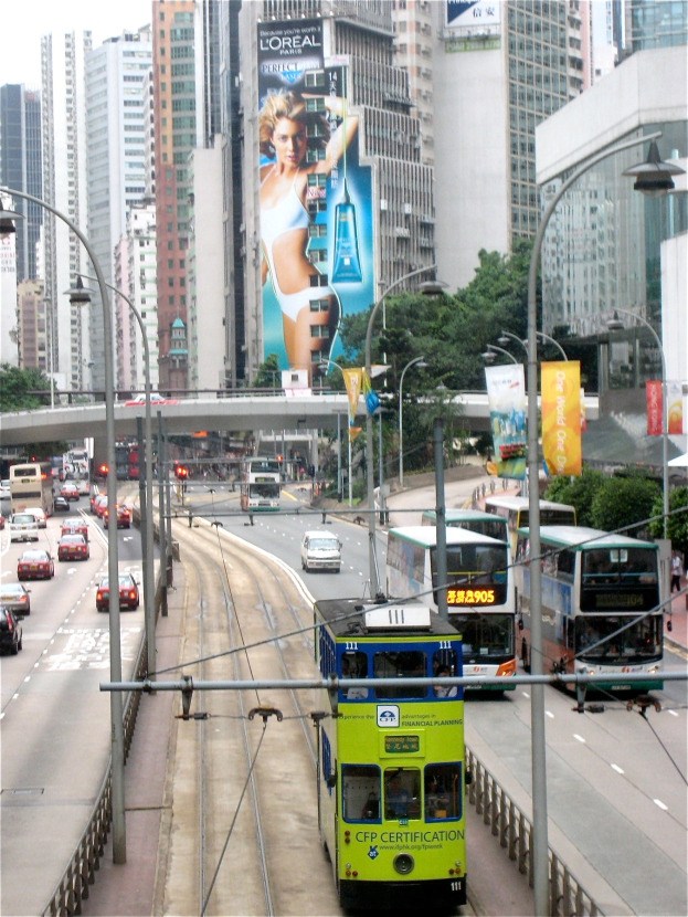 Eine belebte Stadtstraße mit verschiedenen Fahrzeugen, darunter Doppeldeckerbusse, Autos und andere Fahrzeuge, umgeben von Pfählen, Laternen, Schildern, Bannern, Bäumen, einer Brücke und hohen Gebäuden unter einem klaren blauen Himmel, mit Text am unteren Bildrand.