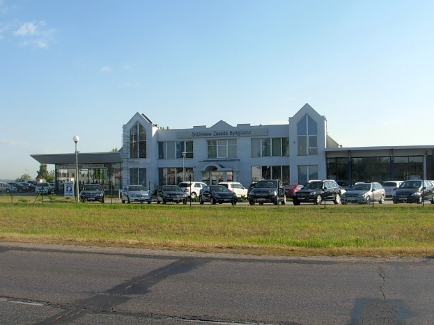Autohaus mit mehreren parkenden Autos, umgeben von Gebäuden, Straßeninfrastruktur, fahrenden Fahrzeugen und Bäumen unter einem bewölkten Himmel.
