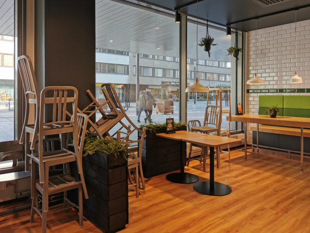 Ein Restaurantinnenraum mit Holztischen und -stühlen, großen Fenstern mit Blick auf eine Straße in Berlin, Deutschland, umgeben von Pflanzen und Dekolicht.
