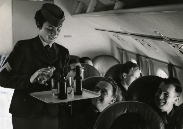 Schwarzes und weißes Bild einer Flugbegleiterin, die Getränke an sitzende Passagiere serviert.