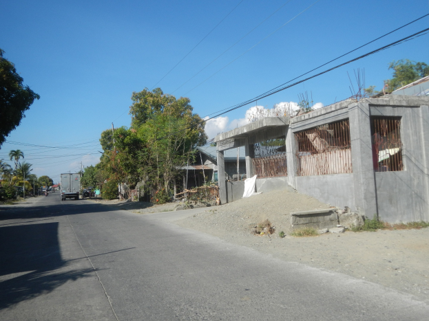 Eine Straße mit einem Gebäude im Bau, fahrende Fahrzeuge, Bäume, Strommasten mit Drähten und einen bewölkten Himmel.